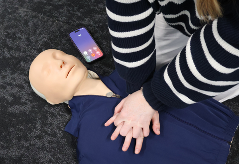 person practicing chest compressions on a training manikin with a phone next to them with 999 dialed