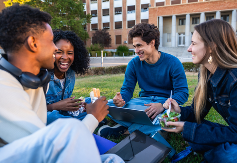 four university students sitting on grass outside a building with their laptops, laughing and chatting