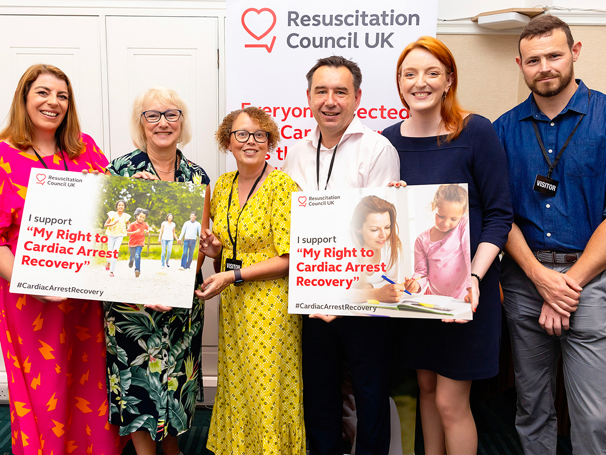 Four women and two men stand facing the camera wearing smart clothing, holding placards that read "My Right to Cardiac Arrest Recovery""