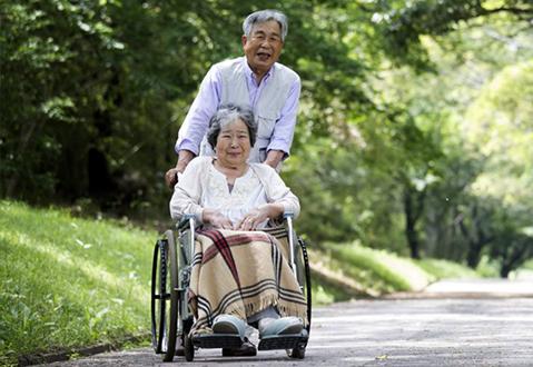 A man and a woman enjoying a walk through a green park, she is using a wheelchair and he is pushing her