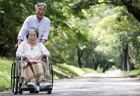 A man and a woman of East Asian descent enjoying a walk through a green park. The woman is being pushed in a wheelchair, and has a blanket over her knees. 