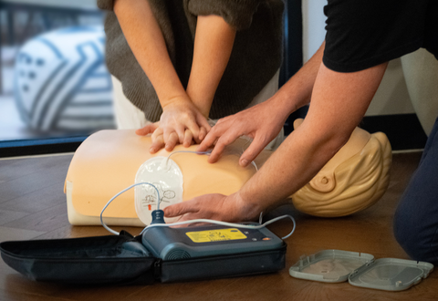 person performing chest compressions on a training manikin while another person attaches defibrillator pads