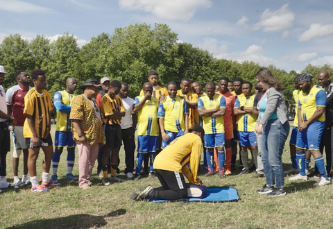 Football team in an area of deprivation, crowded around on a field watching a CPR demonstration