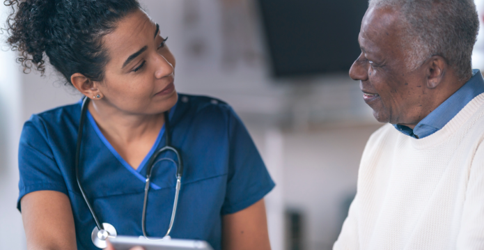 A female healthcare professional in a navy tunic speaking to an older man in a white jumper and shirt.