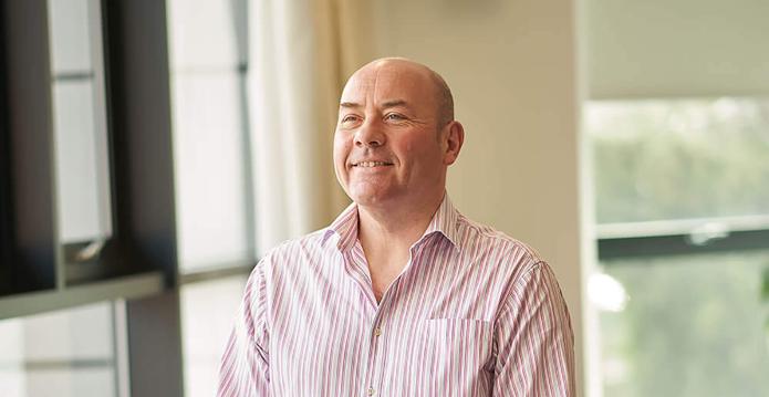 A photo of a tall white smiling man in a striped shirt and black jeans with his hands crossed in front of him.
